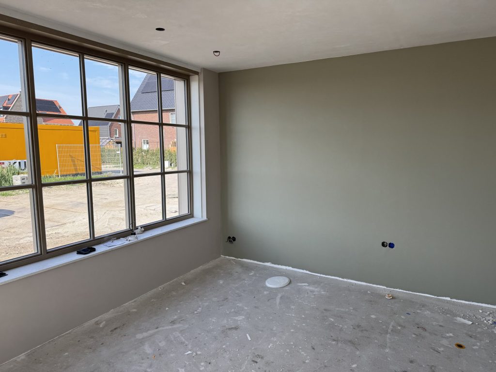 Nieuwbouwkamer met een egaal afgewerkte grijsgroene wand en een groot raam met uitzicht op nieuwe woningen