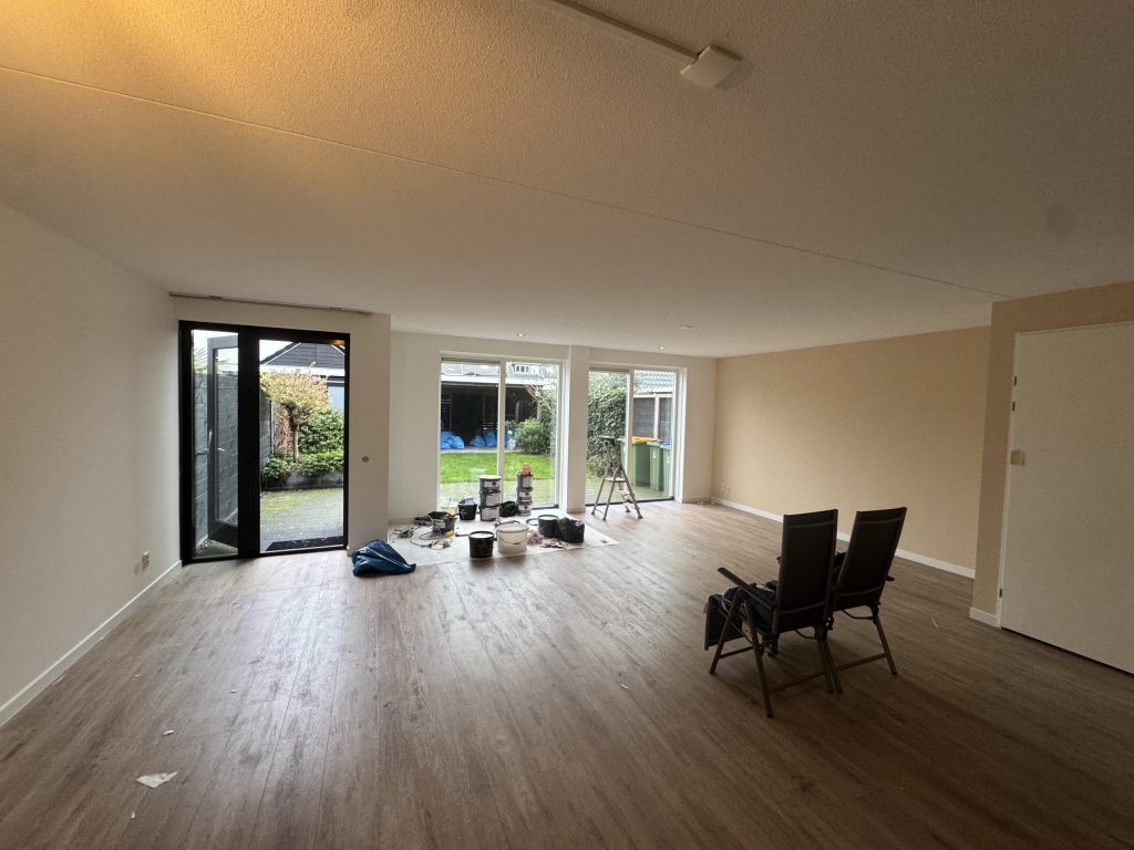 Ruime woonkamer met lichte gesausde wanden, een houten vloer en tuindeuren tijdens de afwerking